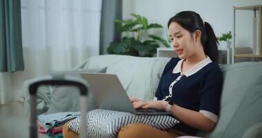 Side view of Asian teenager woman sitting on sofa using laptop for prepare booking hotel and airplane ticket for travel. backpacker travel concept. photo