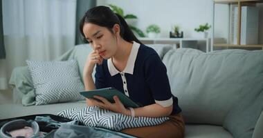 Side view of Asian teenager woman sitting on sofa planning vacation trip and searching information on digital tablet, travel and lifestyle. photo