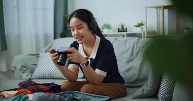 Selective focus of Asian teenager woman wear wireless headphones sitting on sofa look pictures on the camera while packing suitcase luggage in the living room , Preparation travel suitcase at home. photo