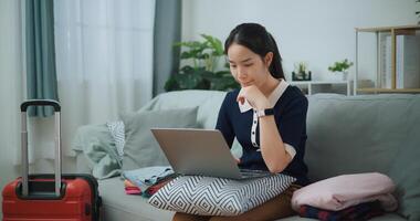 Portrait of Asian teenager woman sitting on sofa using laptop for prepare booking hotel and airplane ticket for travel. backpacker travel concept. photo