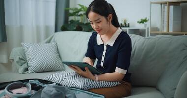 Portrait of Asian teenager woman sitting on sofa planning vacation trip and searching information on digital tablet, travel and lifestyle. photo