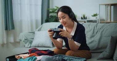 selectivo atención de asiático adolescente mujer vestir inalámbrico auriculares sentado en sofá Mira imágenes en el cámara mientras embalaje maleta equipaje en el vivo habitación , preparación viaje maleta a hogar. foto