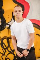 A young man stands in front of a graffiti wall, wearing a white shirt photo