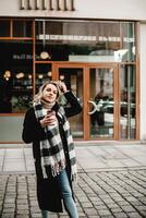 un elegante mujer soportes antes de un edificio, adornado en un negro Saco y tartán bufanda, participación un vibrante rojo café taza. el imagen mezclas urbano Moda con un acogedor café descanso ambiente foto