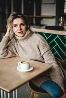 pensativo mujer con café en acogedor cafetería. un mujer en un suéter se sienta a un café mesa, mano en su cabeza, profundo en pensamiento con un taza de café antes de su. foto