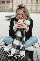 un alegre mujer se sienta en un banco, envuelto en un acogedor bufanda, saboreando un taza de café con un encantador sonrisa. el escena exuda calor y relajación, Perfecto para un tranquilo momento capturar foto