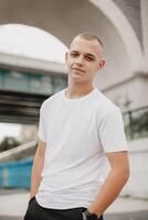 A young man wearing a white shirt stands in front of a bridge photo