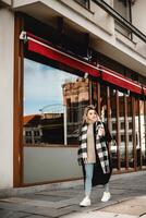 A stylish woman in a black coat and plaid scarf walks gracefully along a city sidewalk, the vibrant red awning adding a pop of color to the urban scene photo