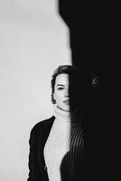 A woman stands before a wall, her gaze fixed on the camera with a serious expression. Clad in a black jacket and white sweater, the image exudes a moody and dramatic ambiance photo