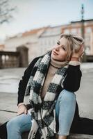 Smiling Woman in Black Coat and Plaid Scarf. Cheerful woman seated on ground, wearing stylish black coat and plaid scarf, tilting her head with a radiant smile, exuding warmth and charm. photo