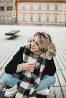 un mujer se sienta en un banco, envuelto en un bufanda, disfrutando un beber. el sereno parque ajuste evoca un sentido de relajación y tranquilidad. acogedor momento en el parque. foto