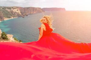 woman red silk dress sits by the ocean with mountains in the background, her dress swaying in the wind. photo