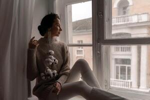 Free time for rest. Profile of a beautiful woman sitting on a white window sill at home, holding a cotton plant in her hands. She is wearing a sweater, leggings and white boots. photo