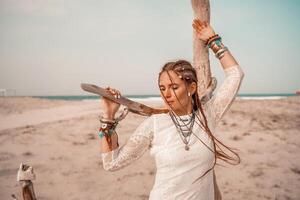 Model in boho style in a white long dress and silver jewelry on the beach. Her hair is braided, and there are many bracelets on her arms. photo