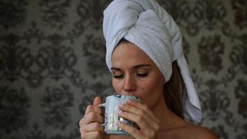 Middle-aged woman looks good with bare shoulders in a white towel on her head holds a cup and drinks coffee or tea against the wall photo