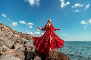 rojo vestir mar mujer. un rubia con fluido pelo en un largo fluido rojo vestir soportes en un rock cerca el mar. viaje concepto, foto sesión a mar