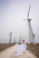 A woman in a white dress is walking down a dirt road in front of a row of wind turbines. photo