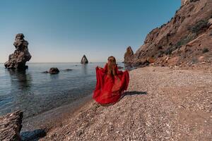 un mujer en un rojo vestir soportes en un playa con un rocoso orilla en el antecedentes. el escena es sereno y pacífico, con el mujer rojo vestir contrastando en contra el natural elementos de el playa. foto
