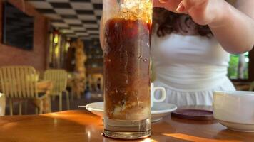 Brown carbonated drink with ice stands on the table in a cafe, hand putting a straw into the drink, background bokeh. Cocktail. High quality 4k footage video