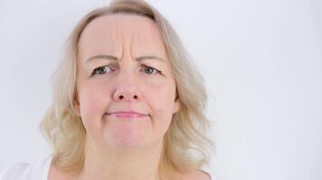 Photo of thoughtful woman bites lower lip, looks with dreamy expression aside, wearing stylish beige hoodie, stands against white wall. Copy space for your text video