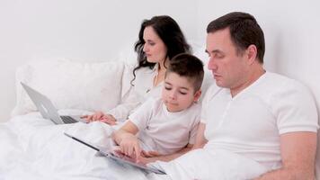 famille, paternité, La technologie et gens concept content père et peu fils avec tablette PC ordinateur séance dans lit à Accueil maman femme travail dans le Contexte en ligne sur ordinateur portable video