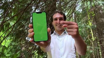 telefone com uma verde tela croma chave dentro a mãos do uma homem ele é em pé dentro a floresta dentro uma branco camisa mostrando a telefone para a Câmera video