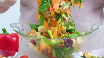 A woman mixes ingredients of vegetable salad slow motion with falling lettuce being mixed in glass plate carrots arugula spinach tomatoes croutons red pepper cucumbers vegetarian mix video