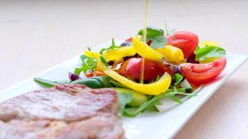 Grüner Salat ist bewässert mit Sonnenblume oder Olive Öl Tomaten Pfeffer Grüns Grüner Salat Blätter Koriander neben Fleisch Schweinefleisch auf ein Weiß rechteckig Teller Nahansicht köstlich Essen Mittagessen gegrillt Fleisch Portion Geschirr video