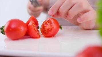 Nahansicht von ein Kinder Hand falsch Schneiden ein Kirsche Tomate Junge oder Mädchen Lernen zu Schnitt Gemüse mit ein Käse Messer Nächster zu Mamas Hand Gemüse Weiß Porzellan Tafel Kochen Lernen zu Koch video