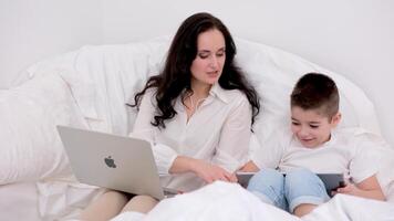 mamá y hijo sentar en un blanco cama en su manos manzana tecnología ordenador portátil macbook tableta ipad y móvil teléfono iphone 13 Pro max Mira a cada otro sonrisa usos el último tecnología amor cuidado riqueza es video