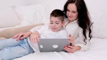 mom and son are lying comfortably on the bed relaxing playing a game on a tablet calling home have gone to another country white clothes black hair love caring family relationships motherhood video