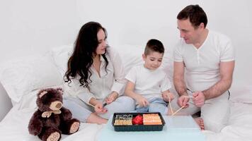 spend time with family in white clothes playing with chopsticks from oriental food wooden sticks drum laugh have fun in bed together eat sushi soft toy brown teddy bear day off dad mom son video