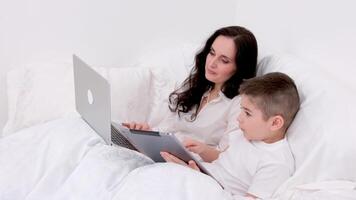 Mama und Sohn unter Decke sind Sitzung auf Sanft Bett im das Hände von Junge Tablette spielen online Freiberufler auf Laptop verbringen Zeit mit Sohn während Arbeit genesen bleibe beim Zuhause Uhr Film online Kommunikation video