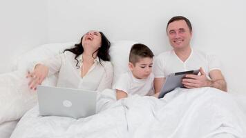 family in bed playing on tablet sincerely laughing having fun spending time together mom father and son dark hair white big duvet family is watching movies with the son on laptop video