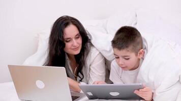 trabalhos às casa conectados mulher debaixo cobertor trabalhando em computador portátil ao lado filho jogando jogos em tábua doente às casa pegue bem família isolamento quarentena branco Folha edredon Preto cabelo mãe e filho video