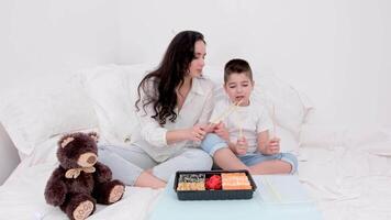 un mujer y su hijo jugar con arroz palos me gusta batería, golpe en el mesa en un suave cama, blanco hojas, acogedor almohadas, comer Sushi en el cama, relajarse, osito de peluche oso juguete, blanco ropa video