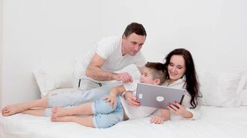 family in white bed with tablet examining game father showing how to press buttons laughing rejoicing child learning new game developing lessons cheerful family love at home spending time together video