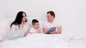 husband wife and son spend time together in bed holiday look at tablet communicate conversation love at home happiness joy success in gadgets white background place for advertising text happy family video