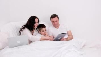 husband wife in bed with little son looking at tablet cartoon interesting game pictures laughing talking communication real people at home love life joyful people space for text ad social relations video