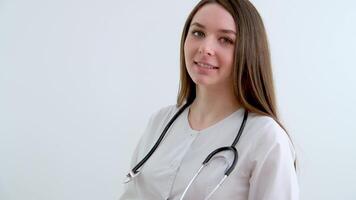 sonrisa médico joven niña en un blanco antecedentes hermosa joven médico enfermero estetoscopio cabeza Disparo en pie en hospital. contento hembra terapeuta, médico, general facultativo mirando a cámara video