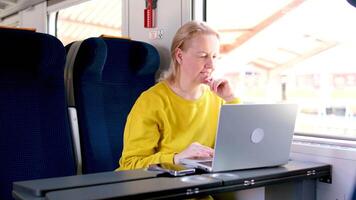 un mujer paseos en un tren un sentado interurbano coche trabajos en un ordenador portátil a un mesa un justa de edad mediana mujer anuncia un teléfono demostración un ordenador portátil riendo espacio para publicidad texto video