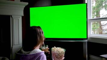 statique trépied coup de Jeune femme en train de regarder film sur la télé avec vert écran et en mangeant pop corn tandis que séance sur canapé. fille à la recherche à préféré télévision spectacle sur chrominance clé afficher dans Accueil vivant chambre. video