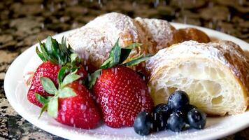 su bianca piatto quattro fragole, fresco frutti di bosco, mirtilli o lokhina e delizioso arioso fresco brioche, uno è tagliare poroso dentro è visibile, cosparso con in polvere zucchero piace neve su marmo tavolo video