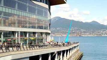 you can see the statue of a drop from the Pacific Ocean restaurants cafes glass office building mountains white clouds people walking along the embankment seagull flying in the sky port Canada Place video