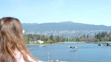 jung Mädchen steht beim Geländer von das Balkon und sieht aus in das Entfernung beim das Berge und Schiffe im das Center von Vancouver tragen Sonnenbrille begradigen ihr Haar lila Farbe im Kleider Ferien Ausflug video