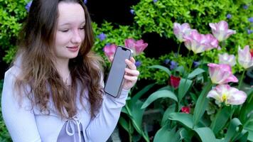 cerca un arbusto con tulipanes, un joven niña muestra un teléfono con un blanco pantalla, sorpresa, ligeramente abierto su boca, Mira a el pantalla, un sitio para publicidad texto, un floreciente jardín parque video