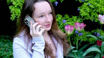 un calma conversacion en el teléfono en el calle un joven niña soportes cerca el arbustos en el parque tulipanes y azul flores en un poción hablando levanta su Cejas empuja su frente hermosa real personas video