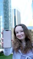 en contra el fondo de un rascacielos en céntrico vancouver, un joven niña muestra un teléfono con un blanco pantalla recomienda un viaje espacio para texto sonrisa lila ropa color adolescente video