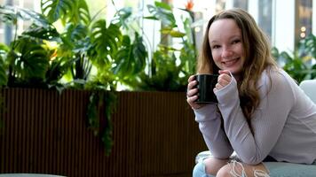 un niña bebidas café con Placer desde un negro taza se sienta cerca el ventana en el salón plantas en el antecedentes rascacielos Rasgado pantalones rubio pelo un sonrisa en su cara un agradable pasatiempo video