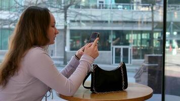 jovem mulher encantadora ligando com o telefone celular enquanto está sentada sozinha na cafeteria durante o tempo livre, mulher atraente com um sorriso fofo conversando com o celular enquanto descansa no café video